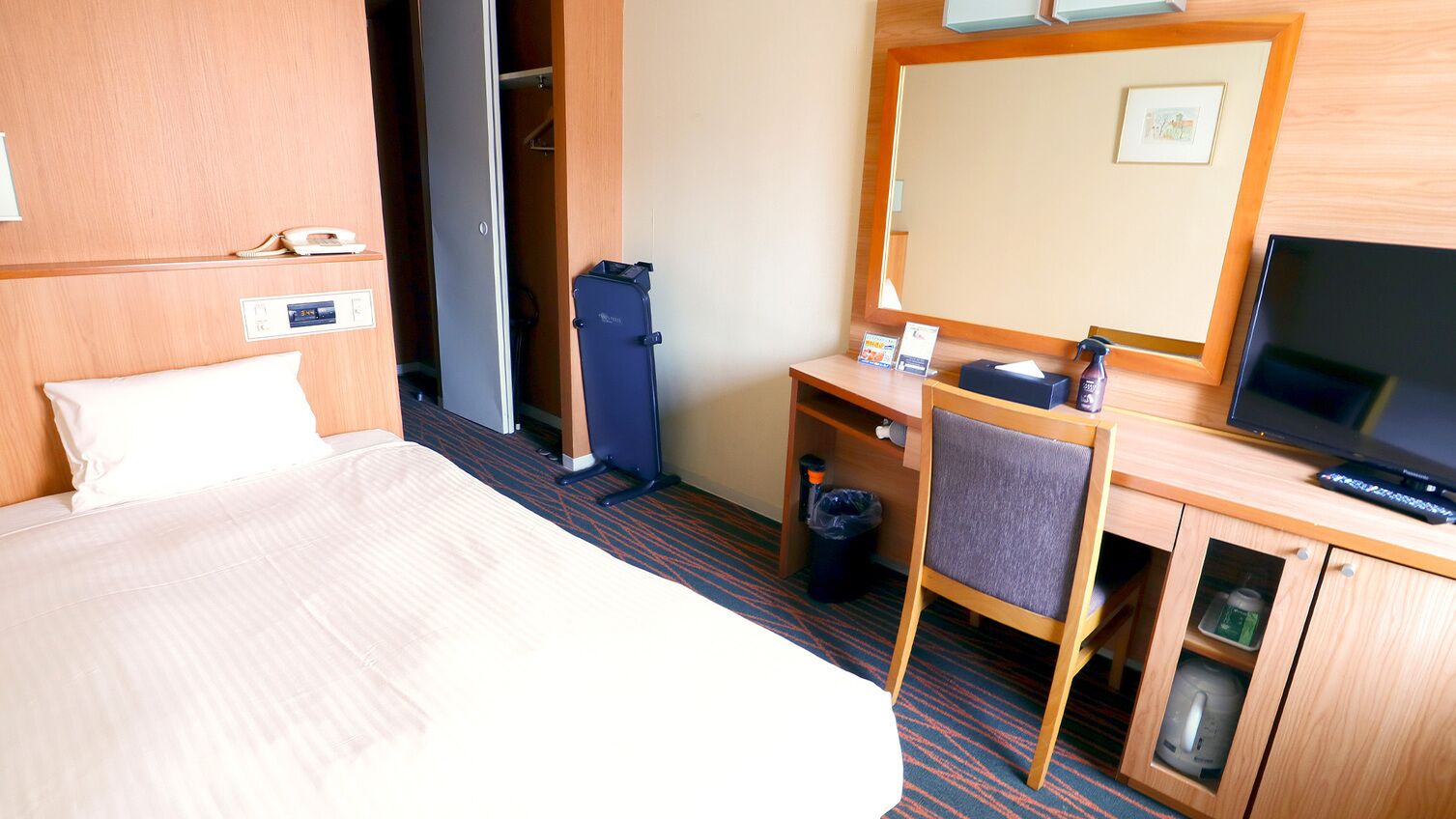 A hotel room with a bed, a desk with a chair, a television, a mirror, a wardrobe, and a small trash bin.