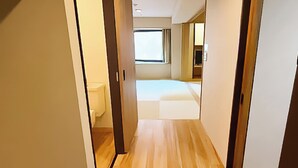 A narrow hallway leading to a bedroom with a large window, wooden flooring, and a visible bathroom.