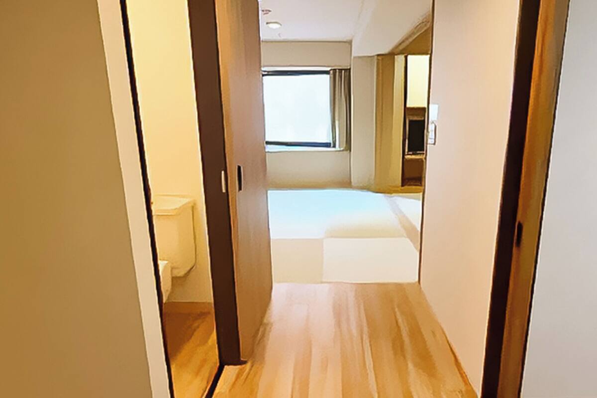 A narrow hallway leading to a bedroom with a large window, wooden flooring, and a visible bathroom.