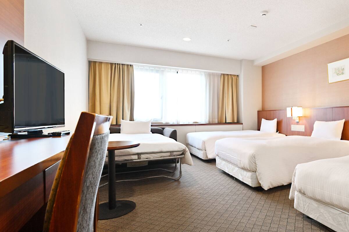 A hotel room with two beds, a desk with a TV, and a window with curtains.