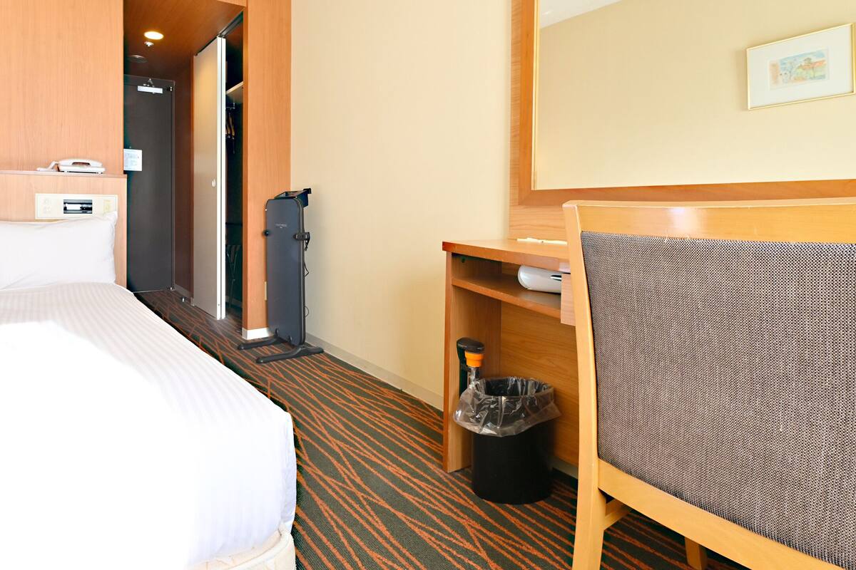 A hotel room with a large bed, a desk with a chair, a trash bin, and a coat rack.