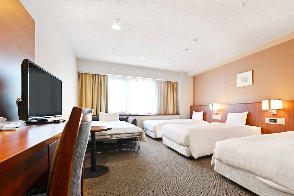 A hotel room with a desk, television, two beds, a chair, and a window with curtains.