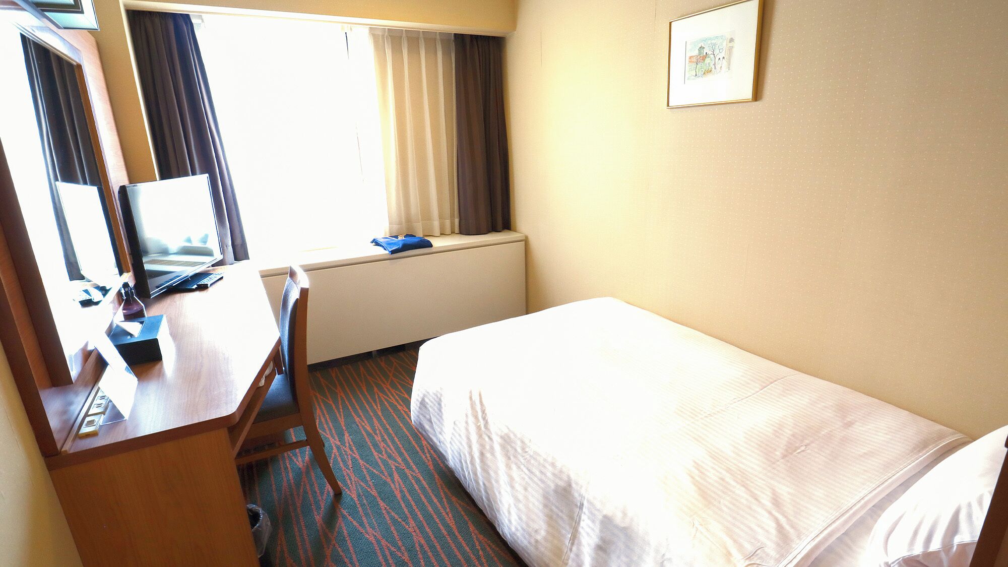 A hotel room with a bed, a desk with a computer, a window with curtains, and a framed picture on the wall.