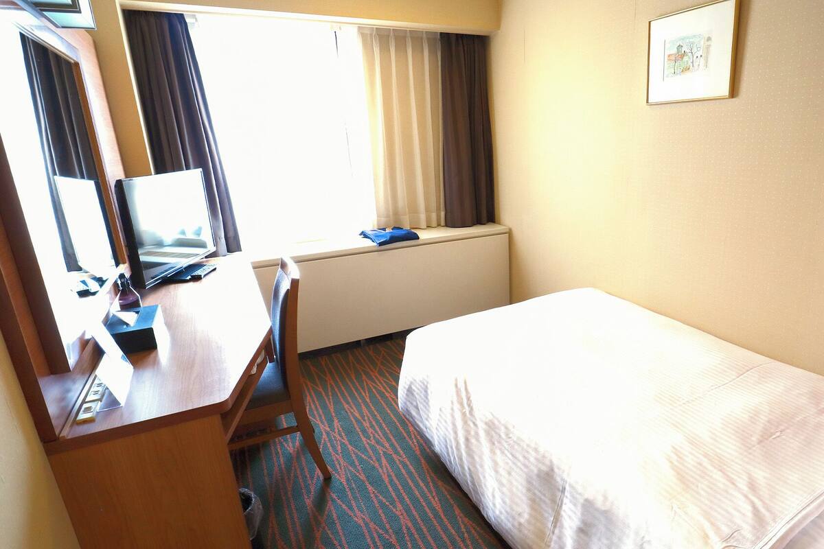 A hotel room with a bed, a desk with a computer, a window with curtains, and a framed picture on the wall.