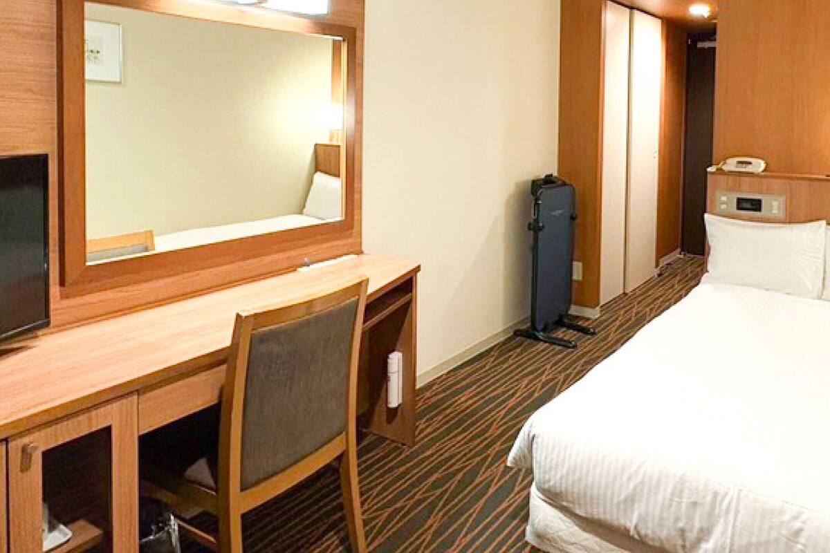A hotel room with a bed, a desk with a chair, a television, a mirror, a wardrobe, and a bathroom.