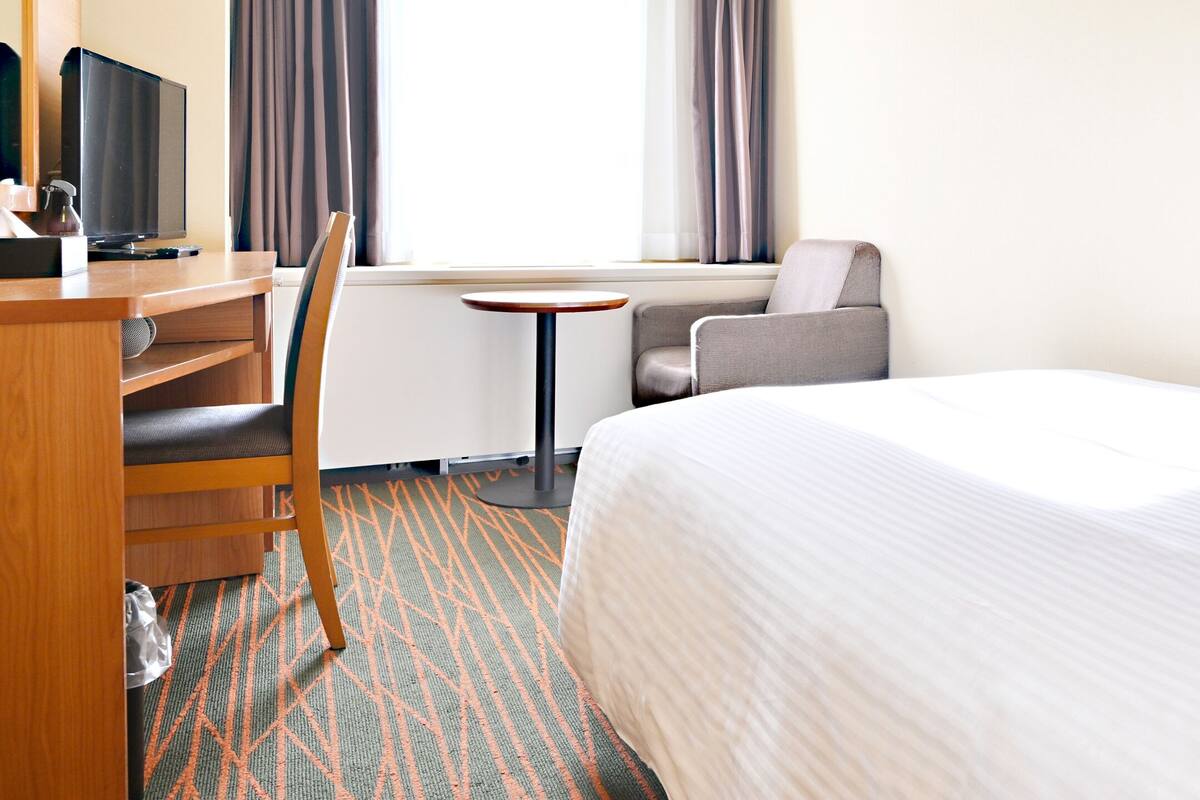 A hotel room with a bed, a desk with a computer, a chair, a small table, and a window with curtains.
