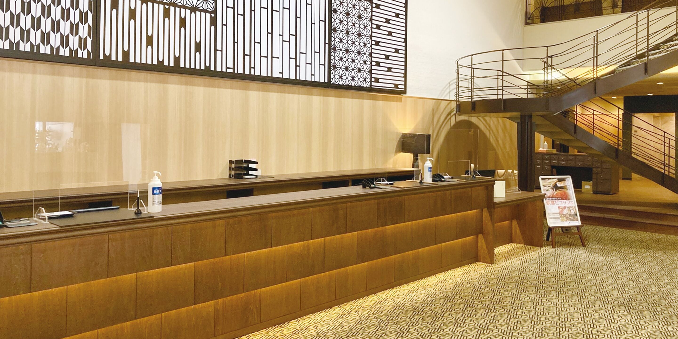 A hotel reception desk with a wooden counter, a digital screen, and a signboard.