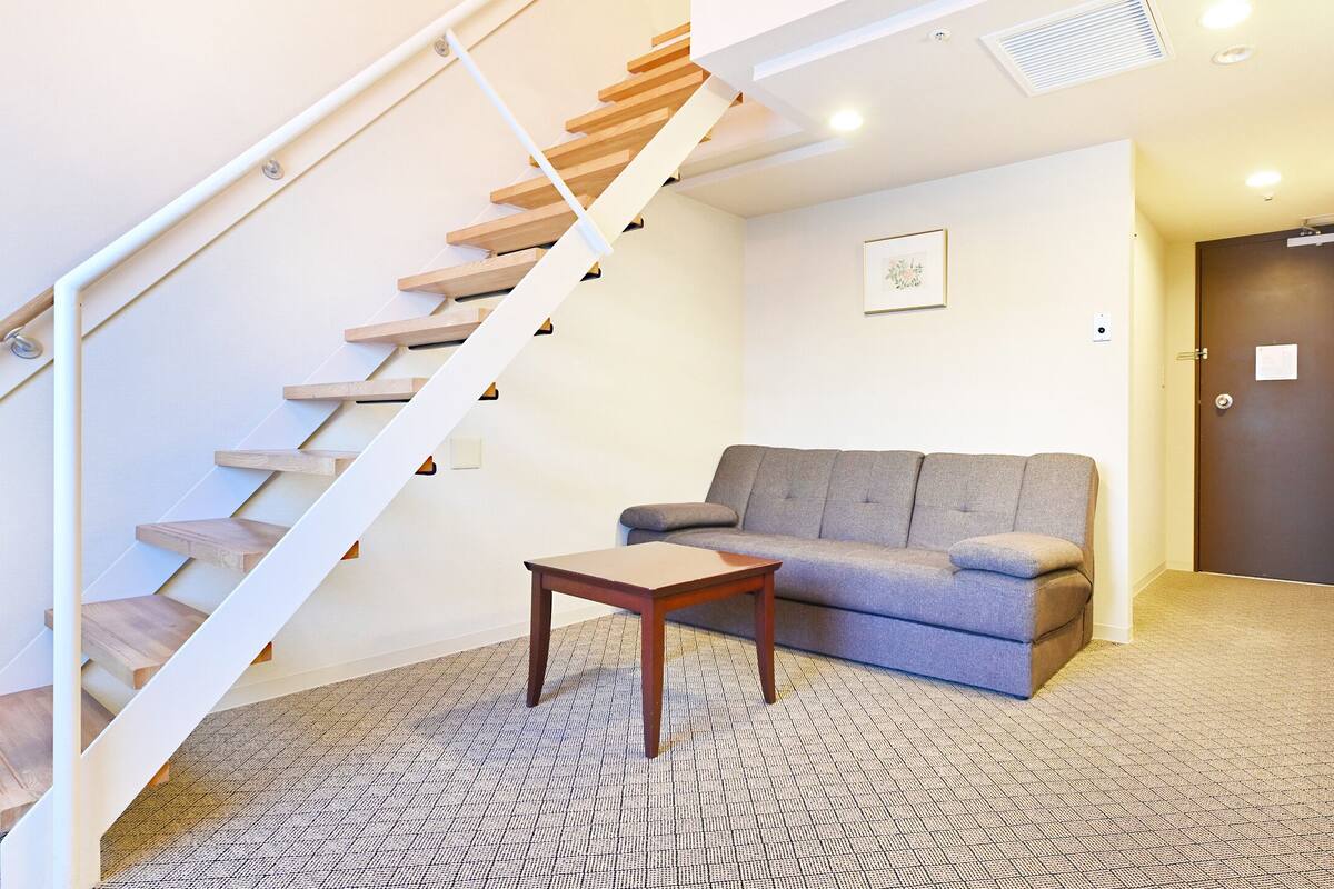 A modern living room with a wooden staircase, grey sofa, small wooden table, and a desk with a chair.