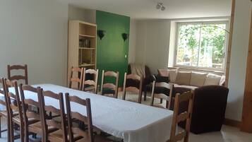 Een eetgedeelte met een lange tafel en stoelen, een boekenkast, een groene accentmuur en een raam met uitzicht op groen.