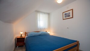 Une chambre avec un lit simple, une table de chevet avec une lampe, une fenêtre avec des rideaux et un tableau encadré au mur.