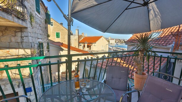 Un balcon avec une table ronde en métal, deux chaises, un pot de fleurs et un grand parasol.