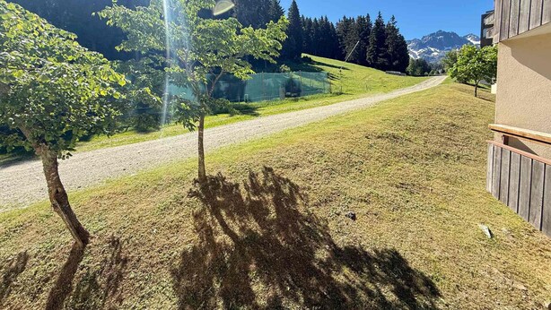 A grassy area with a tree casting a shadow, a gravel path, and a building with a wooden fence. - Cozy 2-room apartment at the foot of the slopes (Les Adrets-Prapoutel)