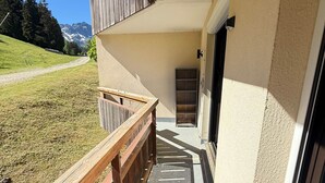 A building with a wooden deck, a door, and a view of a grassy area with mountains in the background. - Cozy 2-room apartment at the foot of the slopes (Les Adrets-Prapoutel)