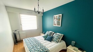 A bedroom with a bed, a nightstand, a dresser, a window with blinds, and a chandelier. - Modern sea-facing apartment with garage and free WiFi (Les Sables-d'Olonne)