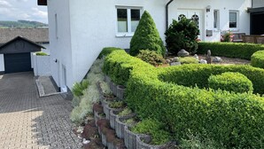 Ett vitt hus med ett tak täckt av tegelpannor, ett garage och en välskött trädgård med häckar och ett litet träd.