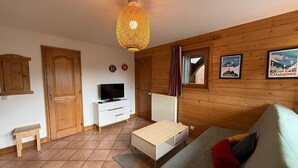 A cozy room with wooden walls, a television on a white cabinet, a bed with a green blanket, and a hanging light fixture. - T3 Apartment in Les Saisies, 6 people, parking included (Les Saisies)