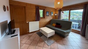 A cozy living room with wooden walls, a sofa, a coffee table, a TV, and a large window offering a scenic view. - T3 Apartment in Les Saisies, 6 people, parking included (Les Saisies)