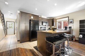 A modern kitchen with dark cabinetry, a black refrigerator, a microwave, and a coffee maker. There is a wooden island with bar stools and a window with blinds. - Townhouse with Free Shuttle to Tremblant Resort (Mont-Tremblant)