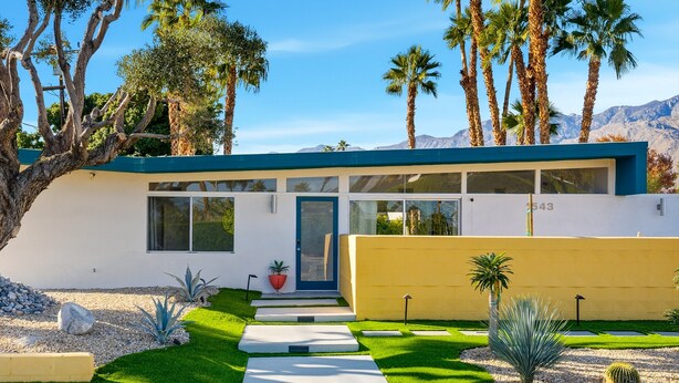 Une maison moderne avec un toit bleu, un mur jaune et un jardin bien entretenu, agrémenté de cactus et de palmiers.