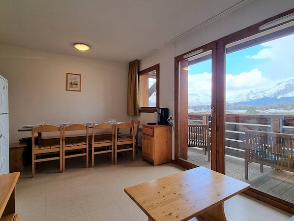 A dining area with wooden chairs and a table, a coffee machine, and a view of a snowy mountain through a glass door.