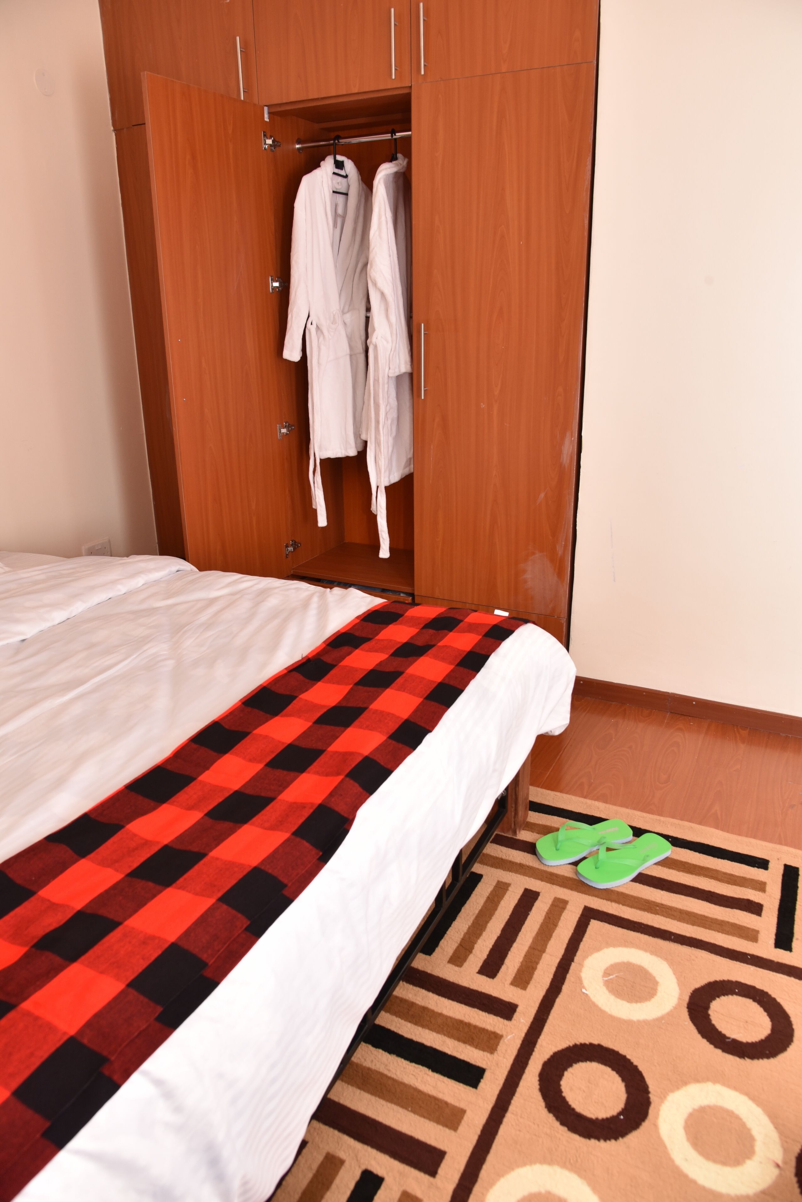 Quarto com cama, manta xadrez, um par de chinelos verdes e guarda-roupa com dois roupões brancos pendurados dentro.