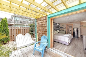 A wooden deck with a swing set and a blue Adirondack chair, leading to a room with a bed and a kitchenette. - Surf's Up at Island Breeze of CB (Carolina Beach)