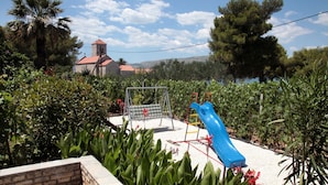 A playground with a blue slide and swings, surrounded by greenery and a building with a red roof in the background. - Large Apartment w. big Terrace, Next to the Beach (Trogir)