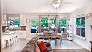 Een ruime woonkamer met een grote bank, een eettafel en stoelen. Er is een keuken met witte kasten en een raam met uitzicht op groen.