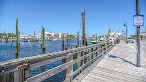 A wooden pier extending into a marina with boats docked, a clear blue sky, and a sign indicating no skateboarding and walk bikes allowed.
