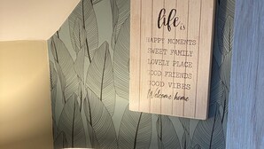 Een lamp op een houten standaard, een tijdschrift op een tafel, en een muur met een bladmotief en een bord met de tekst welkom erop.