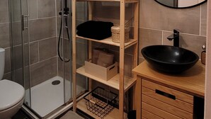 Ein Badezimmer mit einer Duschkabine, einem Holzregal mit Körben und Behältern, einem schwarzen Waschbecken und einem Spiegel an der Wand.
