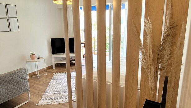 Ein modernes Wohnzimmer mit Holzboden, einem grauen Sofa, einem kleinen weißen Tisch mit einer Pflanze, einem Fernseher und einem großen Fenster mit Blick ins Freie.