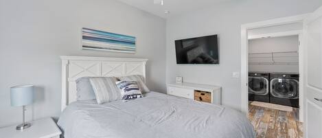 A bedroom with a bed, a nightstand, a ceiling fan, a TV, and a laundry area.