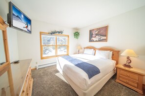 Quarto com cama, mesas de cabeceira, televisão na parede, janela e um quadro.