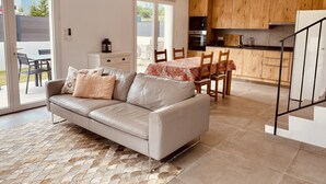 A modern living room with a leather sofa, dining table, and chairs. A kitchen with wooden cabinets and appliances is visible in the background. - Maisonnette de Charme (Arbonne)