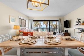 A dining area with a wooden table set for four, a sofa, a TV, and a balcony with palm trees.
