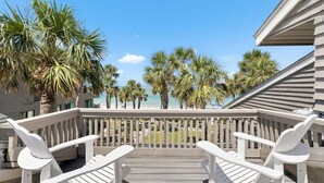A wooden deck with white chairs, overlooking a beach with palm trees and a clear sky.