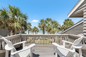 A wooden deck with white chairs, overlooking a beach with palm trees and a clear sky.