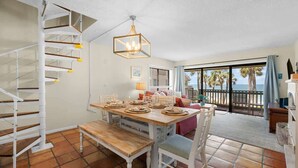 A dining area with a wooden table set for four, a white staircase, and a balcony with a view of palm trees.