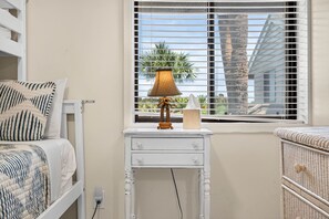 Un dormitorio con cama, mesita de noche, lámpara y vistas a palmeras a través de la ventana.
