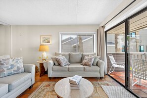 Una sala de estar con sofá, mesa de centro y lámpara. Se ve una ventana con persianas y vistas a edificios.