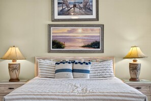 A neatly made bed with striped bedding and two decorative pillows, flanked by two nightstands with lamps and palm tree vases. - Aldea 6 Beachfront Townhome in Belleair Beach (Belleair Beach)