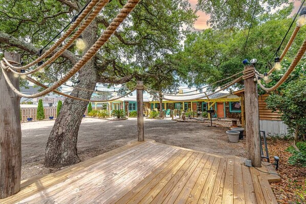 A wooden deck with rope swings, a large tree, and a building with a yellow roof in the background.