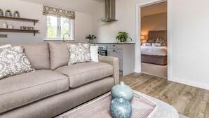 Una sala moderna con un sofá color beige, una zona de cocina con estufa, y un dormitorio visible a través de una puerta abierta.