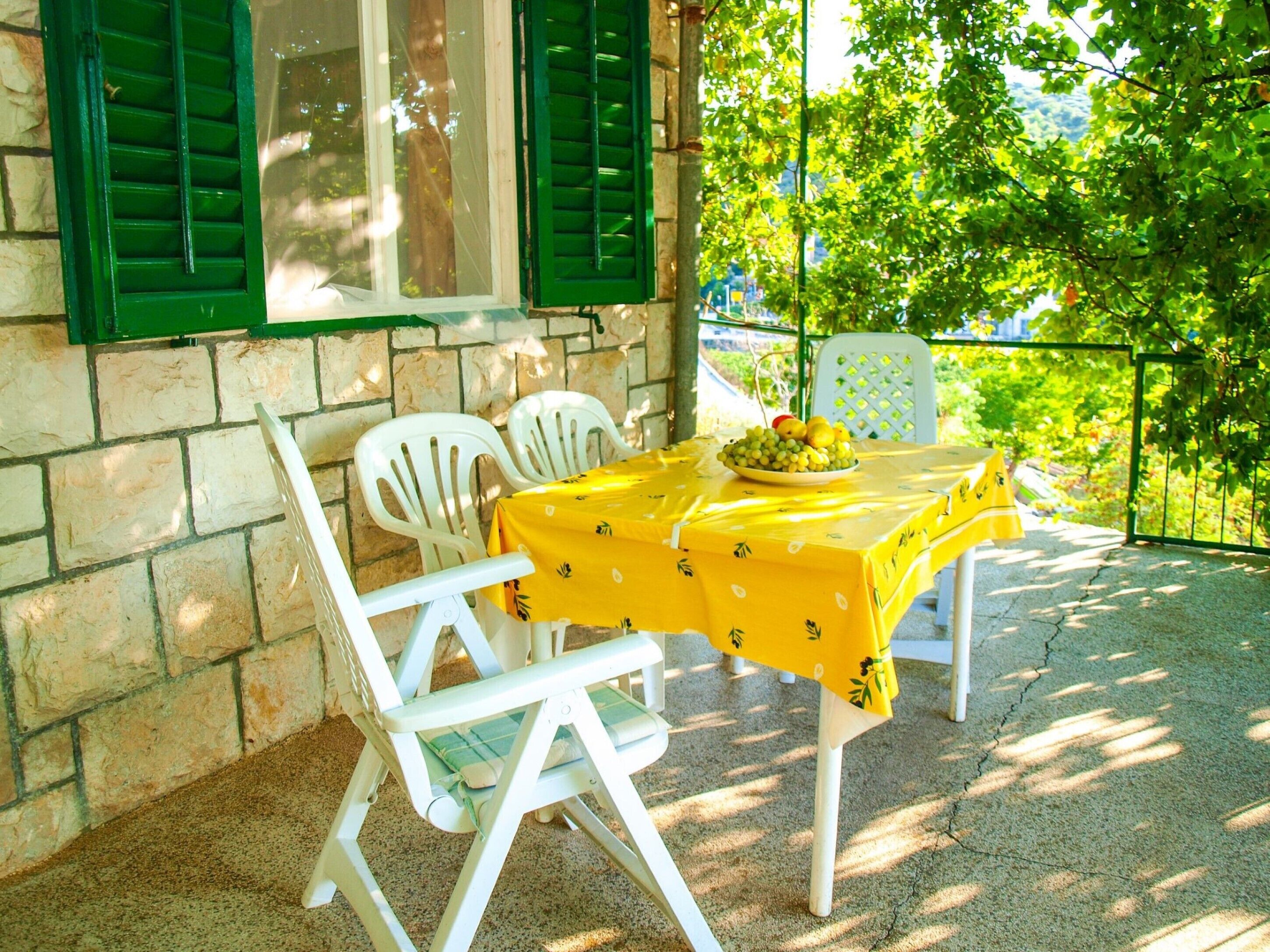 A table with a yellow tablecloth, two white chairs, and a bowl of fruit on a patio with stone walls and green shutters.