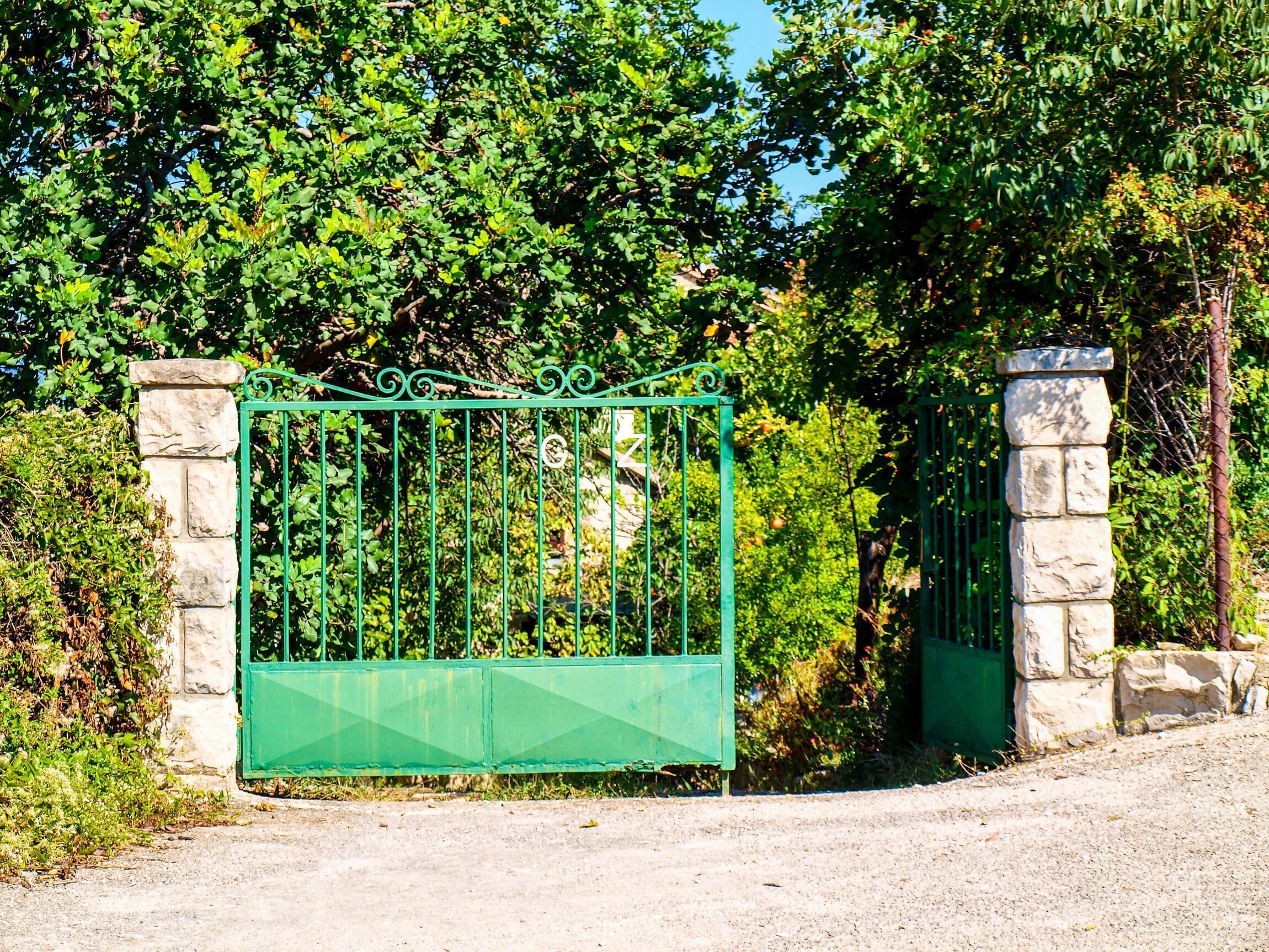 A green metal gate with stone pillars, set in a lush green environment.