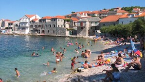 A beach with people swimming and sunbathing, stone buildings with red roofs, and a clear blue sky. - Romantic Apartment With big Garden and Grill (Postira)