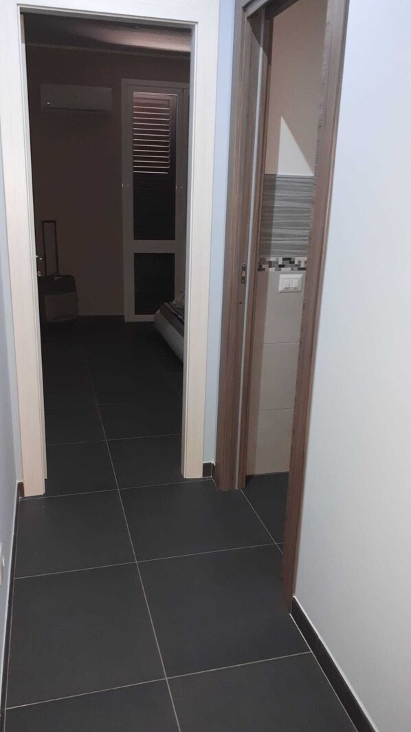 A narrow hallway with dark tiled flooring, a wooden door frame, and a window with blinds.