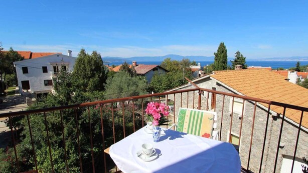 A balcony with a table set for two, overlooking a coastal town with buildings and trees.