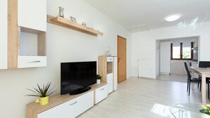 Ein modernes Wohnzimmer mit Flachbildfernseher, Holzausbau, einer kleinen Pflanze und einem Essbereich mit Glastisch und Stühlen.